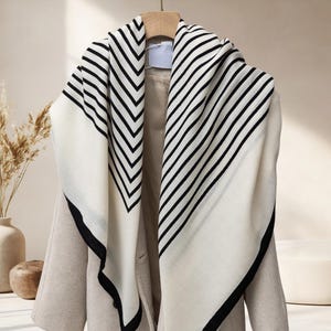 May include: A cream-colored scarf with black stripes and a black border is draped over a beige coat. The scarf has a diagonal striped pattern on one side and a chevron pattern on the other. The coat is hanging on a wooden hanger.