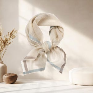 May include: A cream-colored scarf with a knotted design. The scarf has a light blue and gray border. The scarf is made of a soft, flowing material. The scarf is displayed on a white surface.