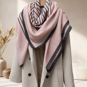 May include: A light pink scarf with dark gray stripes and borders is draped over a light-colored coat. The scarf is made of a soft material and has a simple, elegant design. The coat has a double-breasted design.