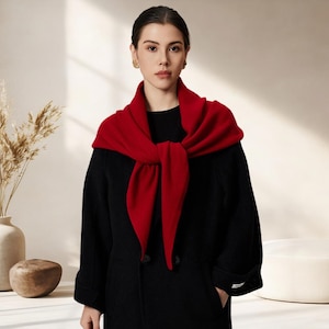 May include: A vibrant red knitted scarf draped over the shoulders of a black coat. The scarf features a soft, textured knit fabric and is tied in a simple knot at the front, contrasting against the solid black long-sleeved coat worn underneath.