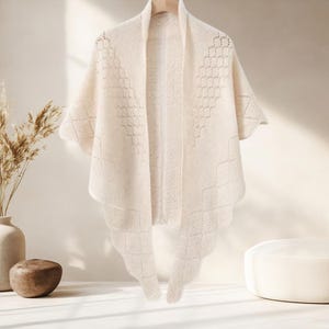 May include: Cream-coloured knitted shawl with a geometric pattern. The shawl is draped over a wooden hanger, showcasing its open weave and scalloped edges. The design features diamond and square patterns.