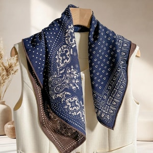 May include: A navy blue scarf featuring a paisley and polka dot design, edged with a brown border. The scarf is displayed on a white vest, suspended from a wooden hanger. The pattern incorporates white floral and geometric shapes.