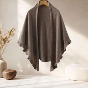 Może przedstawiać: Drapowany szal w kolorze szarobrązowym wisi na drewnianym wieszaku. Szal ma miękki, płynny wygląd z falistym brzegiem. Neutralny kolor i prosty design sugerują wszechstronny dodatek odpowiedni do różnych strojów i okazji.