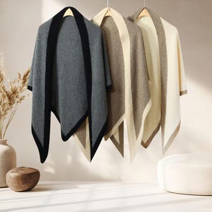 May include: Three shawls in neutral colours hang from wooden hangers. The shawls are grey with black trim, beige with brown trim, and cream with brown trim. The shawls are displayed against a neutral background.
