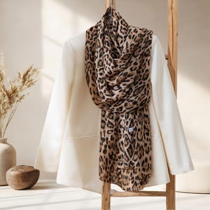 Puede incluir: Una blazer color crema con una bufanda con estampado de leopardo colgada de una escalera de madera. La bufanda presenta un estampado de animales marrón y negro. La escena se desarrolla sobre un fondo neutro con elementos decorativos.