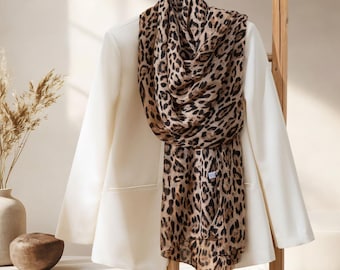 May include: A cream-colored blazer with a leopard print scarf draped over it, hanging on a wooden ladder. The scarf features a brown and black animal print. The scene is set against a neutral background with decorative elements.