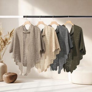 May include: Four open-front shawls in neutral colours hang on a rod. The shawls are beige, cream, grey, and olive green. Each shawl has a scalloped hem and a diamond-shaped openwork design. The shawls are displayed on wooden hangers.