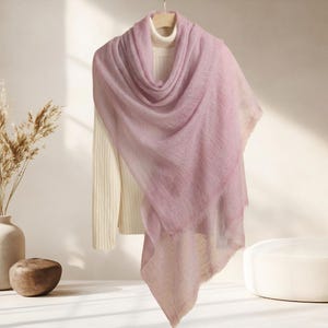 May include: A light purple scarf draped over a cream-colored turtleneck sweater. The scarf has a soft, textured appearance and is hanging on a wooden hanger. The background is a neutral color.