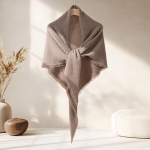 May include: A taupe-colored shawl with a knotted front, fringed edges, and a triangular shape. The shawl is hanging on a wooden hanger against a neutral background, showcasing its soft texture and drape.