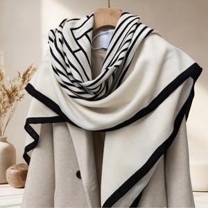 May include: A cream-colored scarf with black geometric patterns and a black border is draped over a beige coat. The scarf is hanging on a wooden hanger. The background includes a vase with dried plants.