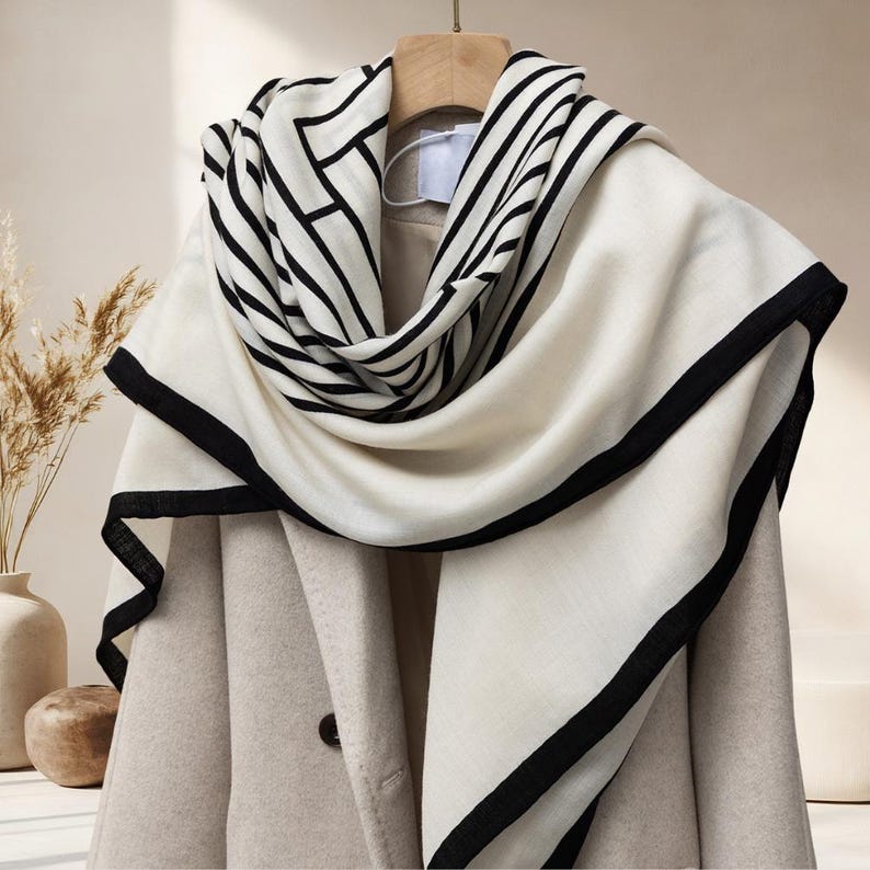 May include: A cream-colored scarf with black geometric patterns and a black border is draped over a beige coat. The scarf is hanging on a wooden hanger. The background includes a vase with dried plants.
