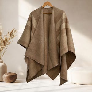 Puede incluir: Un poncho marrón con un sutil patrón de rayas horizontales colgado de una percha de madera. La prenda presenta un diseño frontal abierto con paneles de tela drapeados que caen en pliegues suaves, creando una apariencia en capas con tonos marrones.