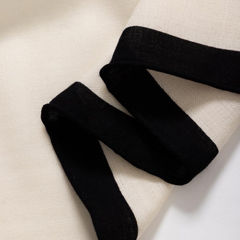 May include: Close-up of a cream-colored fabric with a black border. The fabric has a subtle herringbone weave pattern. The black border is thick and wraps around the fabric.