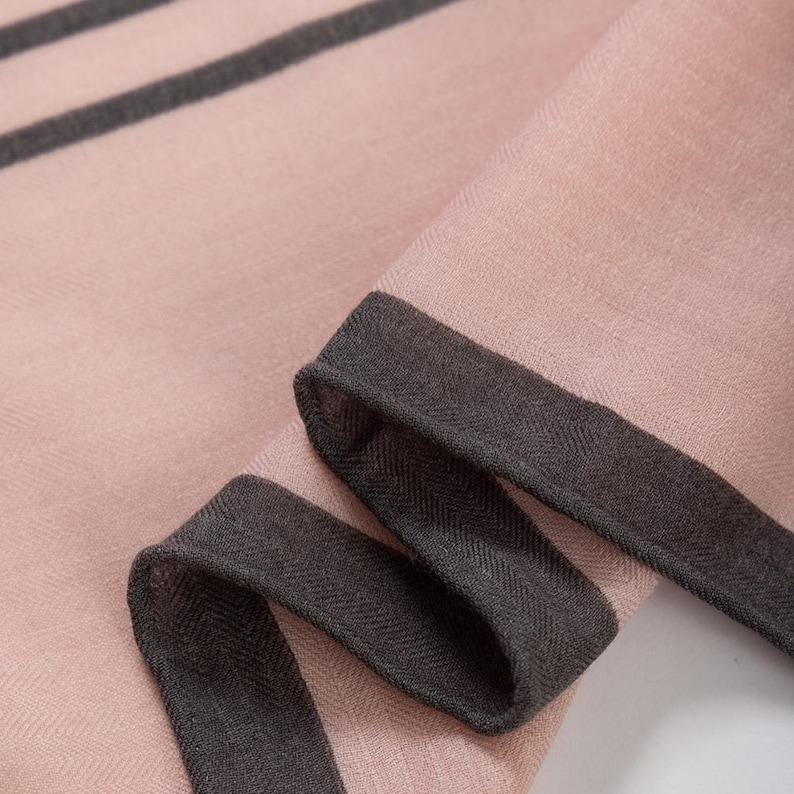 May include: Close-up of a pink scarf with dark gray trim and stripes. The scarf is folded, showing the texture and detail of the fabric. The trim is a darker shade of gray, creating a contrast with the pink background.