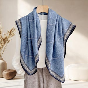 Puede incluir: Bufanda azul con un detallado estampado de cachemira y un borde azul oscuro, drapeada sobre una parte superior blanca. La bufanda se exhibe en una percha de madera. El fondo incluye un jarrón con plantas secas y una pared de color neutro.