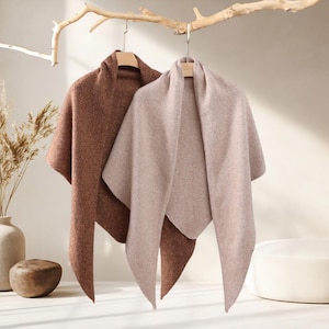 May include: Two triangular shawls hanging on wooden hangers. One shawl is a deep, textured brown, and the other is a light, soft beige. Both garments feature a simple, draped design with pointed ends, showcasing a knit fabric texture throughout.