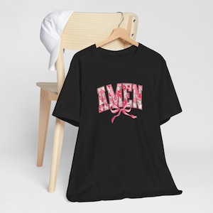 May include: Black t-shirt with the word "AMEN" in a floral pattern with a pink bow. A white baseball cap rests on the back of a wooden chair. The shirt is hanging on a wooden hanger.