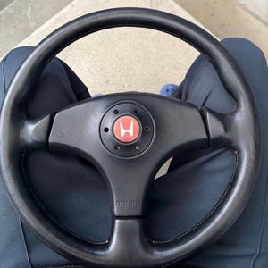 Puede incluir: Un volante de cuero negro con un diseño de tres radios y un botón de bocina circular central. El botón de la bocina muestra un emblema circular rojo con un logotipo de Honda blanco. La palabra MOMO está grabada en el radio inferior del volante.