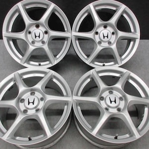 May include: A set of four silver-colored alloy wheels, each featuring a five-spoke design and a central hub cap displaying the black Honda logo. These circular automotive rims are arranged in a two-by-two grid pattern against a neutral background.