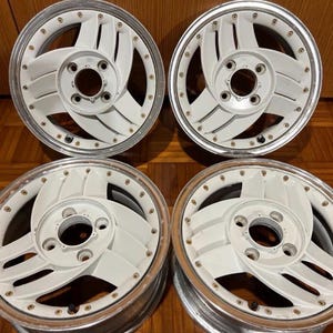 May include: A set of four white automotive wheels featuring a five-spoke turbine design with a polished silver outer rim. Each wheel includes a central hub with five lug nut holes and a series of small decorative bolts along the inner edge.