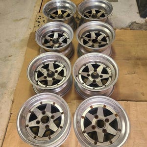 May include: A set of eight silver and dark gray metal automotive wheels arranged in four rows of two. Each wheel features a six-spoke star design with a central hub cap and a polished outer rim, showing signs of wear and oxidation.