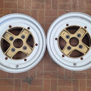 May include: Two circular automotive wheel rims featuring a white outer barrel and a four-spoke gold-toned center hub. Each rim has a central circular opening surrounded by four lug nut holes and a series of smaller holes along the inner perimeter.