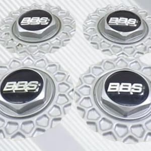 May include: A set of four circular silver wheel center caps featuring a hexagonal center nut design and a black circular inlay with the silver BBS logo text. The outer edge of each cap has a repeating honeycomb pattern in a metallic silver finish.