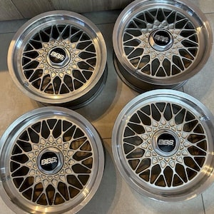 May include: A set of four silver BBS alloy wheels featuring a multi-spoke mesh design with a polished outer rim and a central black cap displaying the white BBS logo. These circular automotive wheels have a metallic finish and a complex geometric pattern.