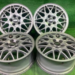 May include: A set of four silver-coloured BBS alloy wheels featuring a multi-spoke cross-mesh design. Each wheel has a central hub cap with the black and silver BBS logo clearly visible. The wheels are arranged in a two-by-two grid formation.