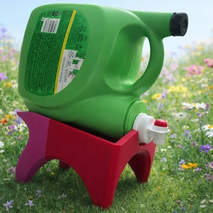 Puede incluir: Un recipiente de plástico verde con asa y tapón negro, colocado sobre un soporte rojo y rosa. El recipiente tiene un grifo blanco con una palanca roja. La escena está ambientada en un campo de flores silvestres.