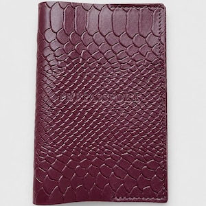 Pode incluir: Uma capa de passaporte em tom bordô profundo com um padrão de pele de cobra texturizado em relevo na superfície. A palavra PETPASSPORT está gravada em letras maiúsculas no centro da capa frontal, que possui bordas costuradas na lateral.