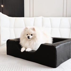 May include: A small, fluffy white dog with pointed ears and a thick coat resting inside a rectangular black pet bed. The bed features a textured, faux-leather finish with high, supportive sides and a low front opening for easy access.