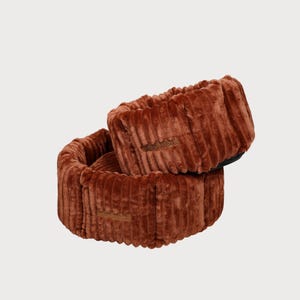 May include: Two rust-colored, corduroy-textured pet beds stacked on top of each other. Each bed has a rectangular shape with rounded corners and a small, brown label with text. The beds are designed for pets.