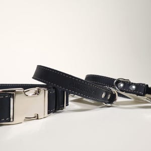Puede incluir: Primer plano de dos collares de cuero negro para perros con hebillas y anillas de metal plateado. Los collares tienen costuras blancas y la marca "POWGARGA" est&aacute; grabada en el cuero.
