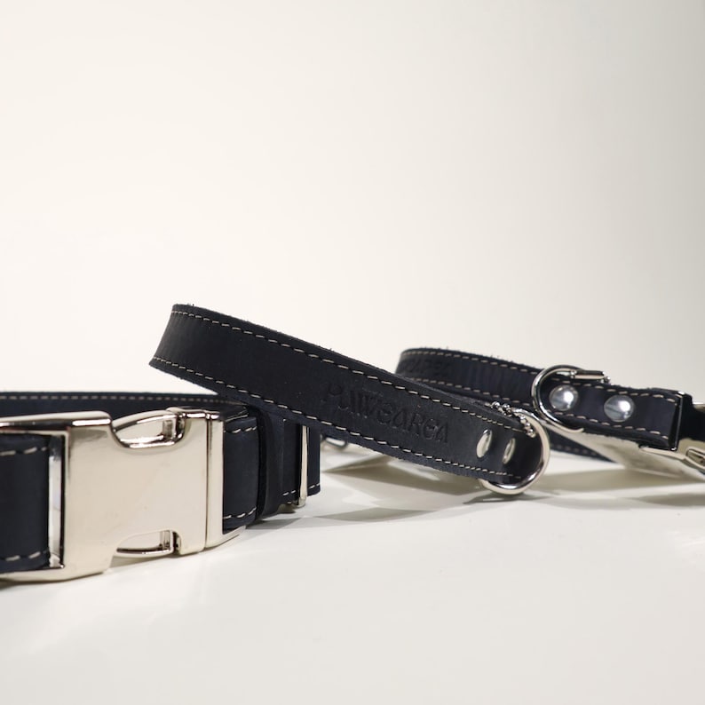 Puede incluir: Primer plano de dos collares de cuero negro para perros con hebillas y anillas de metal plateado. Los collares tienen costuras blancas y la marca "POWGARGA" est&aacute; grabada en el cuero.