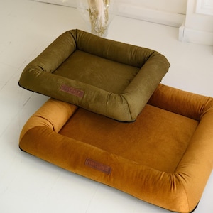 May include: Two rectangular pet beds in olive green and golden brown. The beds have a plush, cushioned design with raised sides and a low entry point. Each bed features a small, rectangular label with text. The beds are designed for comfort and support for small to medium-sized dogs.