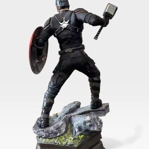 K&ouml;nnte beinhalten: Schwarze Captain America-Figur mit Schild und Hammer. Die Figur steht auf einem felsigen Sockel mit gr&uuml;nem Moos und einem dunklen rechteckigen St&auml;nder. Die Figur tr&auml;gt einen schwarzen Anzug und Helm.