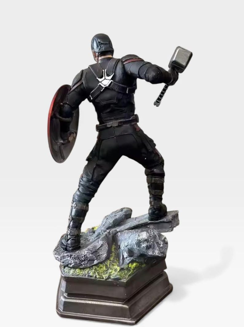 K&ouml;nnte beinhalten: Schwarze Captain America-Figur mit Schild und Hammer. Die Figur steht auf einem felsigen Sockel mit gr&uuml;nem Moos und einem dunklen rechteckigen St&auml;nder. Die Figur tr&auml;gt einen schwarzen Anzug und Helm.