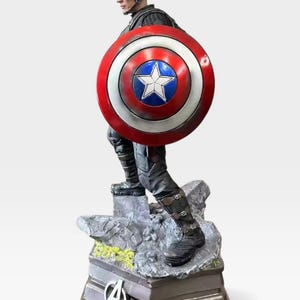 K&ouml;nnte beinhalten: Eine Captain America-Figur, die die Figur in einer dynamischen Pose zeigt, die ihren Schild h&auml;lt. Der Schild hat ein rot-wei&szlig;-blaues Design mit einem wei&szlig;en Stern. Der Sockel der Figur zeigt das Avengers-Logo.
