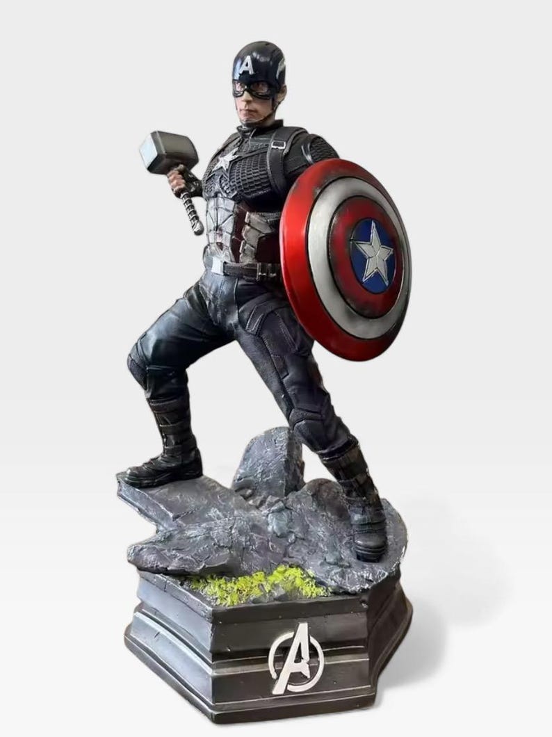 K&ouml;nnte beinhalten: Eine Captain America-Figur, die einen Hammer und einen Schild h&auml;lt. Die Figur befindet sich in einer dynamischen Pose auf einem felsigen Sockel, der sich auf einer schwarzen Plattform mit dem Avengers-Logo befindet. Der Schild ist rot, wei&szlig; und blau mit einem wei&szlig;en Stern. Die Figur tr&auml;gt einen blauen Helm.