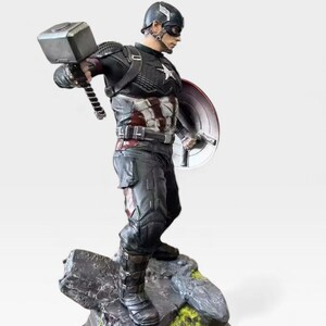K&ouml;nnte beinhalten: Eine Captain America-Figur, die einen Hammer und einen Schild h&auml;lt. Die Figur befindet sich in einer dynamischen Pose und tr&auml;gt einen schwarz-silbernen Anzug mit einem wei&szlig;en Stern auf der Brust. Der Sockel ist ein felsiges Gel&auml;nde.