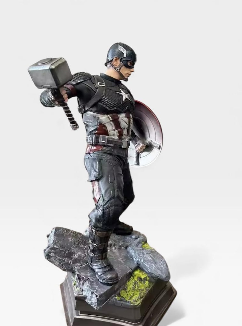 K&ouml;nnte beinhalten: Eine Captain America-Figur, die einen Hammer und einen Schild h&auml;lt. Die Figur befindet sich in einer dynamischen Pose und tr&auml;gt einen schwarz-silbernen Anzug mit einem wei&szlig;en Stern auf der Brust. Der Sockel ist ein felsiges Gel&auml;nde.