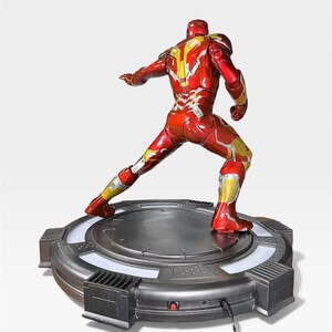 May include: A red and gold Iron Man action figure in a dynamic pose, standing on a circular, metallic base with a glowing white light. The figure's armor features intricate detailing and a glossy finish, showcasing the iconic superhero.