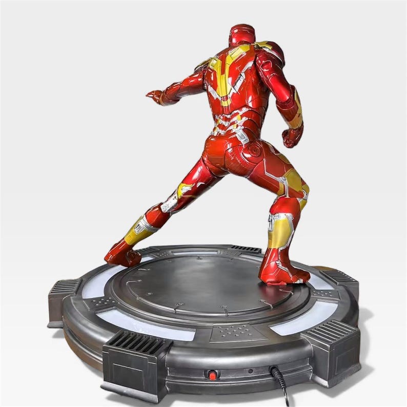 May include: A red and gold Iron Man action figure in a dynamic pose, standing on a circular, metallic base with a glowing white light. The figure's armor features intricate detailing and a glossy finish, showcasing the iconic superhero.