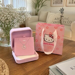 Puede incluir: Un collar de tono plateado con un colgante de Hello Kitty, exhibido en una tarjeta de joyería de terciopelo rosa claro dentro de una caja de terciopelo abierta a juego. Al lado hay una bolsa de regalo de papel rosa con caras de Hello Kitty y flores.