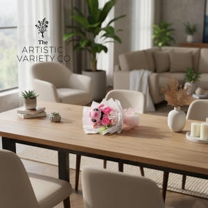 May include: A wooden dining table with a bouquet of pink and white flowers and a stuffed animal. The table also has a small succulent plant, books, and a decorative object. The text "The Artistic Variety Co" is visible.