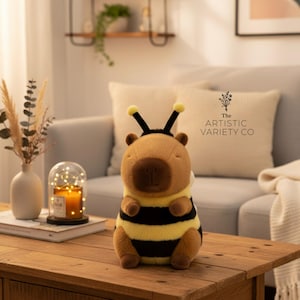 Puede incluir: Un peluche con forma de abeja con cabeza marrón, antenas negras y cuerpo a rayas amarillas y negras. El peluche está sobre una mesa de madera. Una vela encendida en una cúpula de cristal y un jarrón con flores secas también están sobre la mesa.