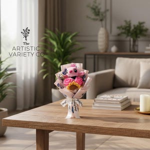 May include: A bouquet with a pink plush character, pink roses, and a yellow sunflower, wrapped in patterned paper and tied with a white ribbon. The bouquet is on a wooden table. The text "The Artistic Variety Co" is visible.