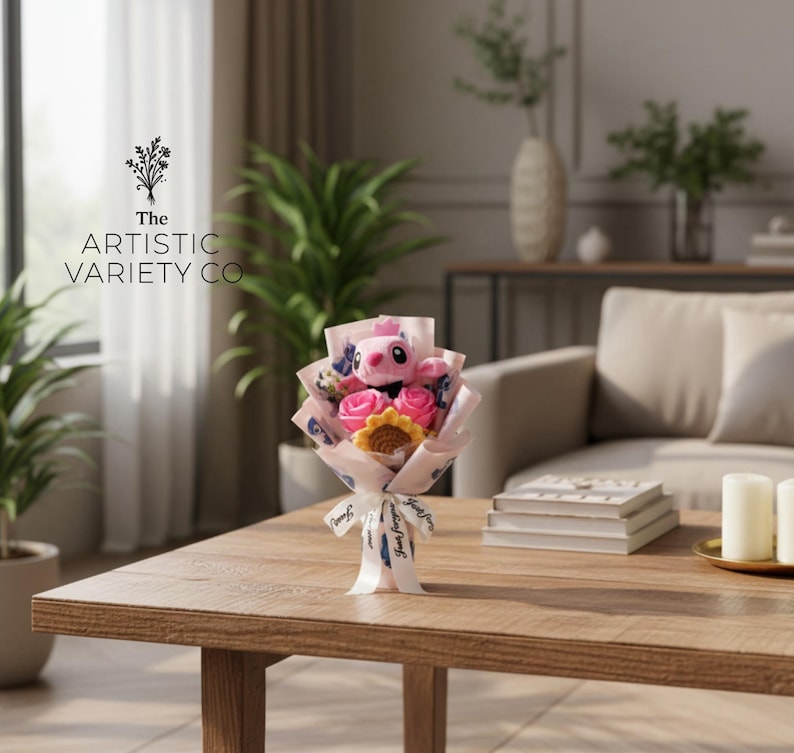 May include: A bouquet with a pink plush character, pink roses, and a yellow sunflower, wrapped in patterned paper and tied with a white ribbon. The bouquet is on a wooden table. The text "The Artistic Variety Co" is visible.