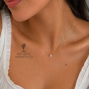 Puede incluir: Un collar de cadena dorada fina con un colgante de piedra transparente de talla brillante redonda. La pieza de joyería se muestra con el texto The Artistic Variety Co y un pequeño icono floral impreso a la izquierda de la cadena del collar.